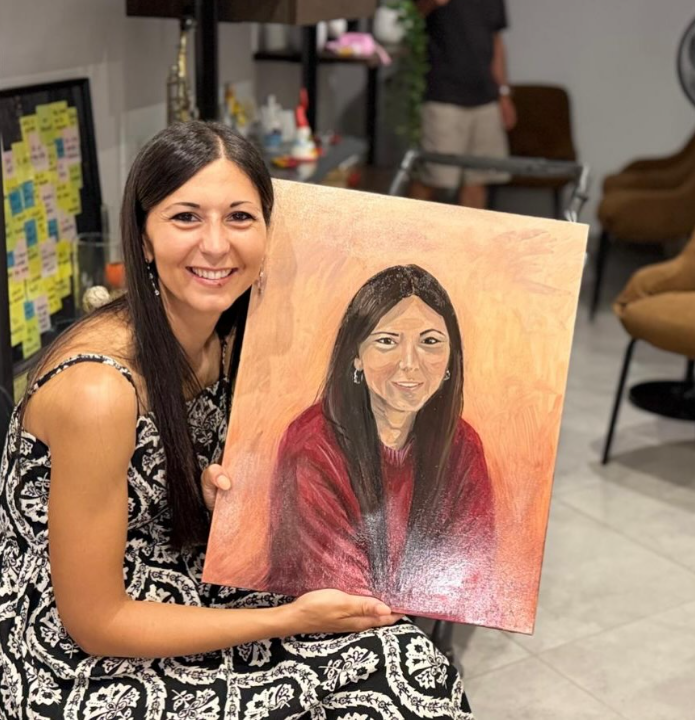 Una donna sorridente con capelli lunghi e vestito scuro a fiori tiene in mano il dipinto che la ritrae con un maglione rosso